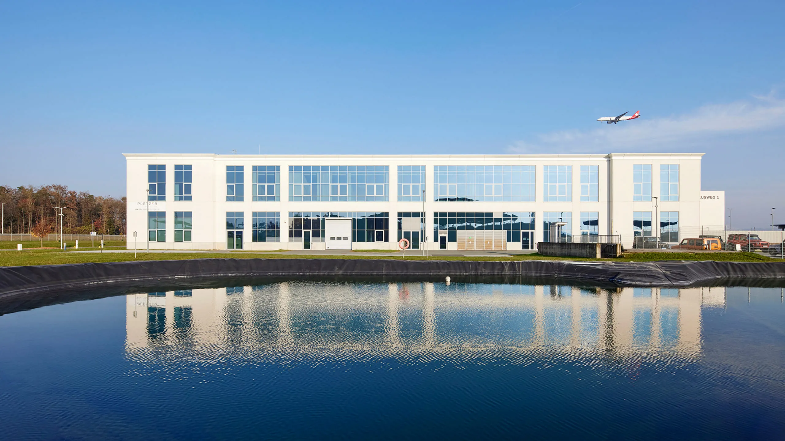 Modernes Bürogebäude von Pletzer Immobilien mit Glasfassade an einem Teich, Flugzeug im Hintergrund.